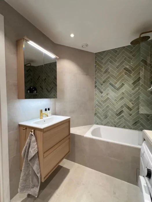 Stilren badrumsrenovering på Östermalm med träkommod, spegelskåp och grönt kakel i fiskbensmönster som fondvägg.